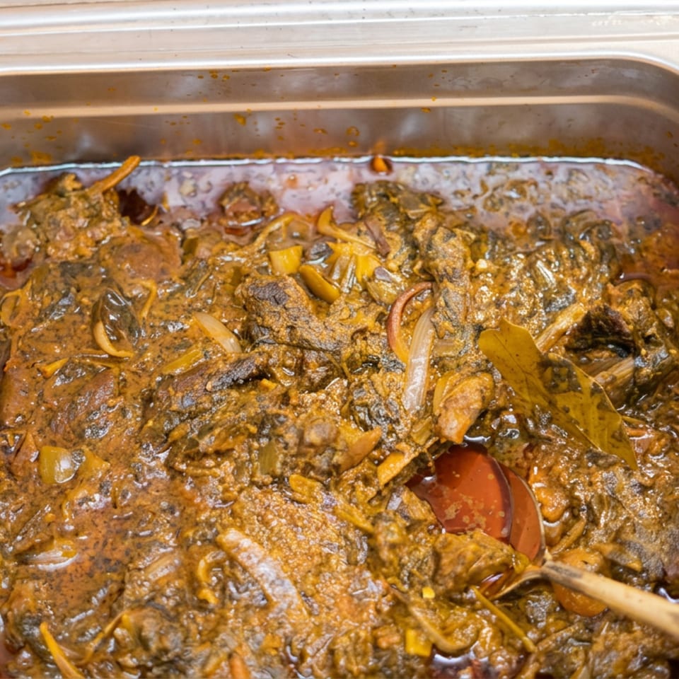 A stainless-steel catering tray holds rich brown stew with sliced onions, peppers, and chunks of meat.